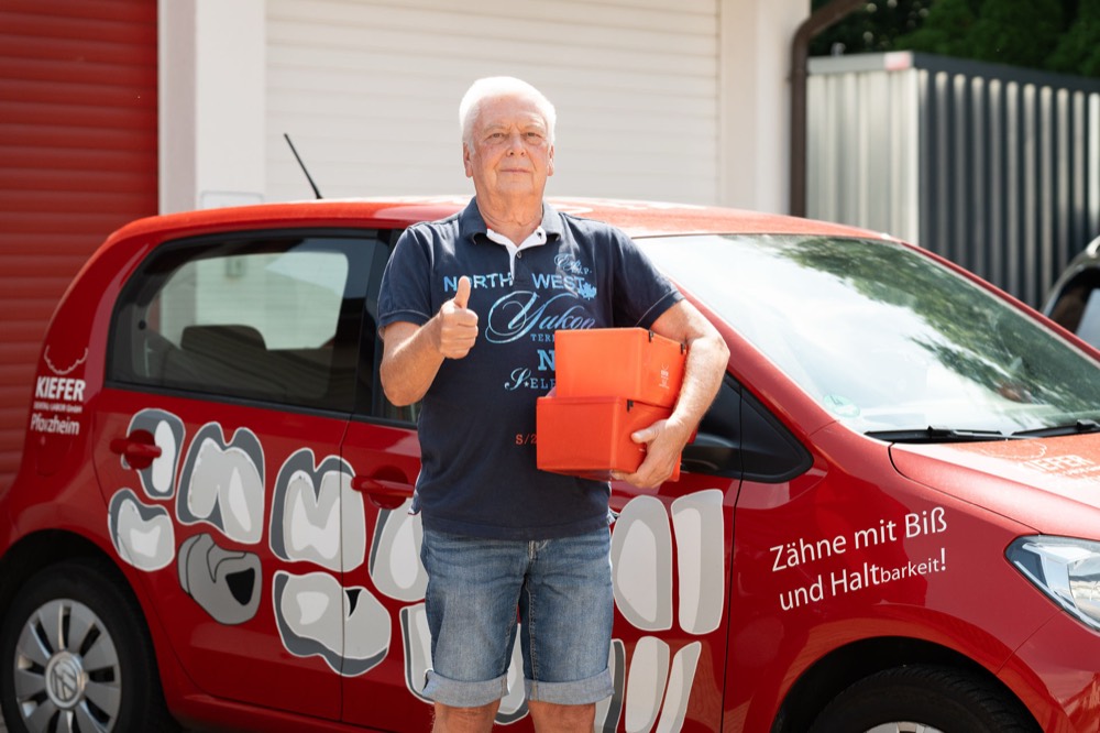 Fahrer des Dentallabors Kiefer mit Transportboxen am roten Fahrdienst-Fahrzeug